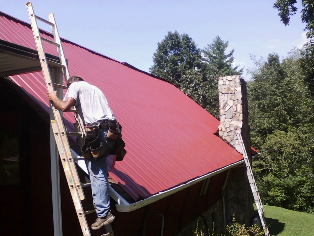 Roofer on a ladder beside a red metal roof and stone chimney — Soffit & Fascia Repair by Roofing Brothers in Rosholt